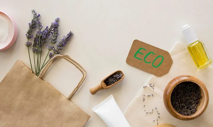 A flat lay of eco-friendly products including lavender, a brown paper bag, a tube, a jar of oil, and a wooden bowl of seeds.